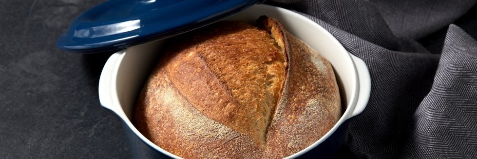 Auf einer dunklen Tischplatte steht ein blau glasierter runder Topf mit einem knusprigen Brot darin. Der Topfdeckel lehnt am Topf und ein graues Geschirrtuch liegt daneben.