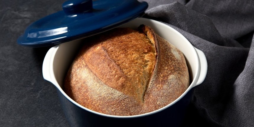 Auf einer dunklen Tischplatte steht ein blau glasierter runder Topf mit einem knusprigen Brot darin. Der Topfdeckel lehnt am Topf und ein graues Geschirrtuch liegt daneben.. Artikel "Brotbacken im Topf" öffnen.