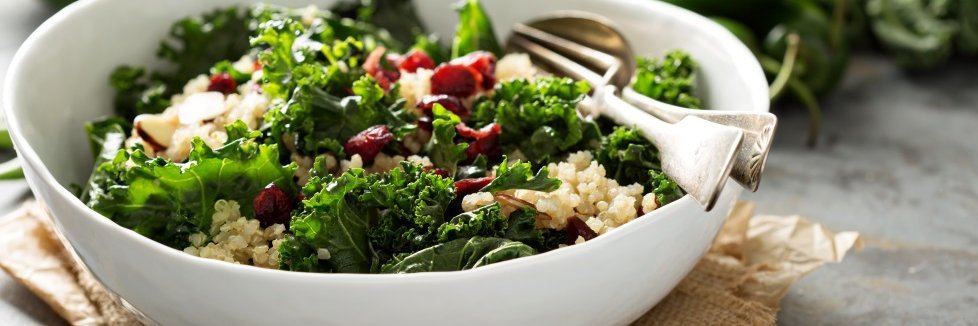 Auf einem dunklen Tisch steht eine weiße Schale mit einem Salat aus Grünkohl, Quinoa, Mandeln und getrockneten Früchten sowie Salatbesteck. Im Hintergrund liegen Grünkohlblätter.