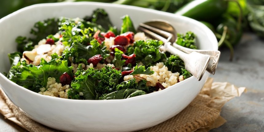 Auf einem dunklen Tisch steht eine weiße Schale mit einem Salat aus Grünkohl, Quinoa, Mandeln und getrockneten Früchten sowie Salatbesteck. Im Hintergrund liegen Grünkohlblätter.. Artikel "Blattkohl ist Gemüse des Jahres" öffnen.