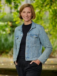 Eine blonde Frau in Jeansjacke vor Hintergrund mit grünen Bäumen.