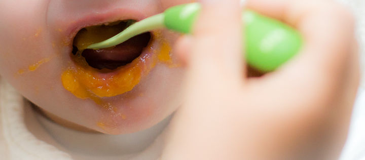 Grüner Löffel füttert Karottenbrei in verschmierten Babymund.