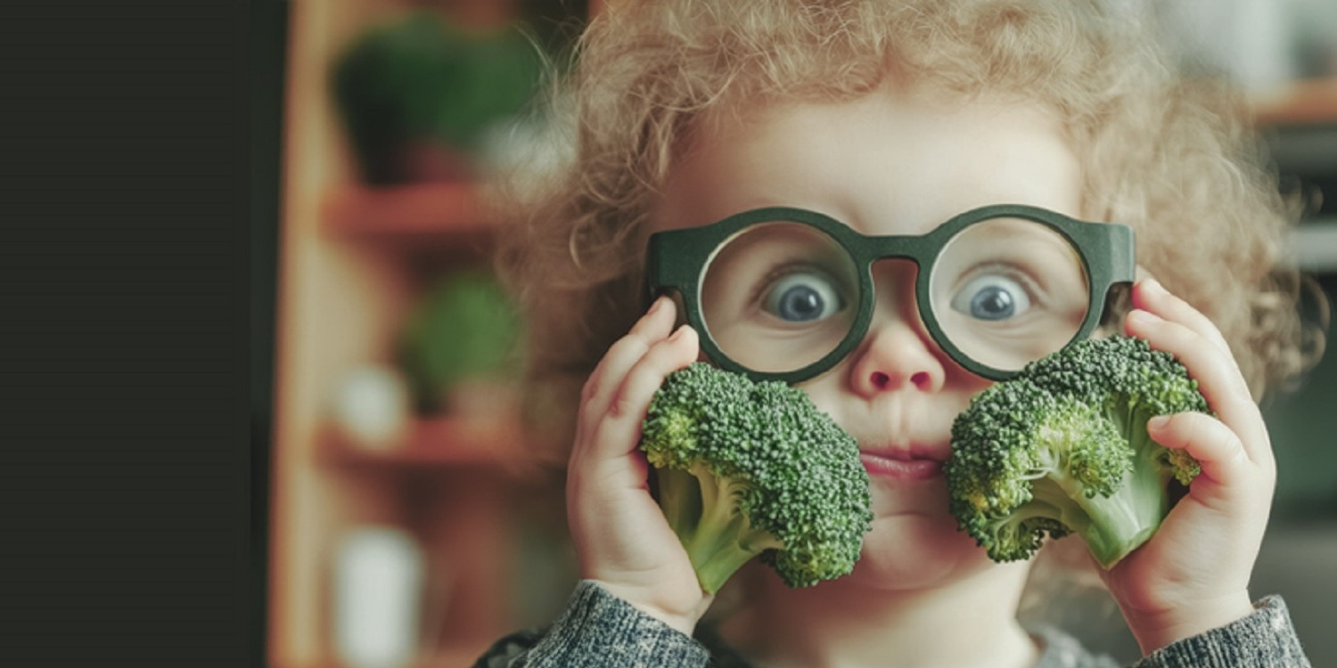 Ein Junge mit dunklem Lockenkopf und einer großen Brille hält sich zwei Brokkoli-Röschen rechts und links an die Wangen.. Artikel "Ernährungsbildung ist das Fundament – nicht die alleinige Lösung" öffnen.