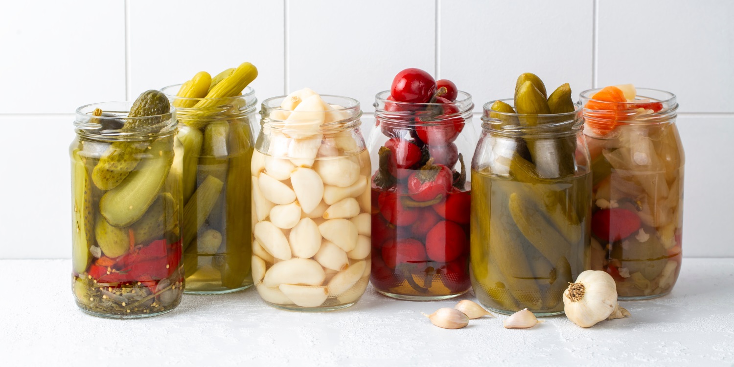 Vor einem weiß gefliesten Hintergrund stehen sechs geöffnete Schraubgläser mit verschiedenen Einlegegurken, Peperoni, Knoblauchzehen und Mixed Pickles.. Artikel "„Mixed Pickles“ selbst zubereiten" öffnen.
