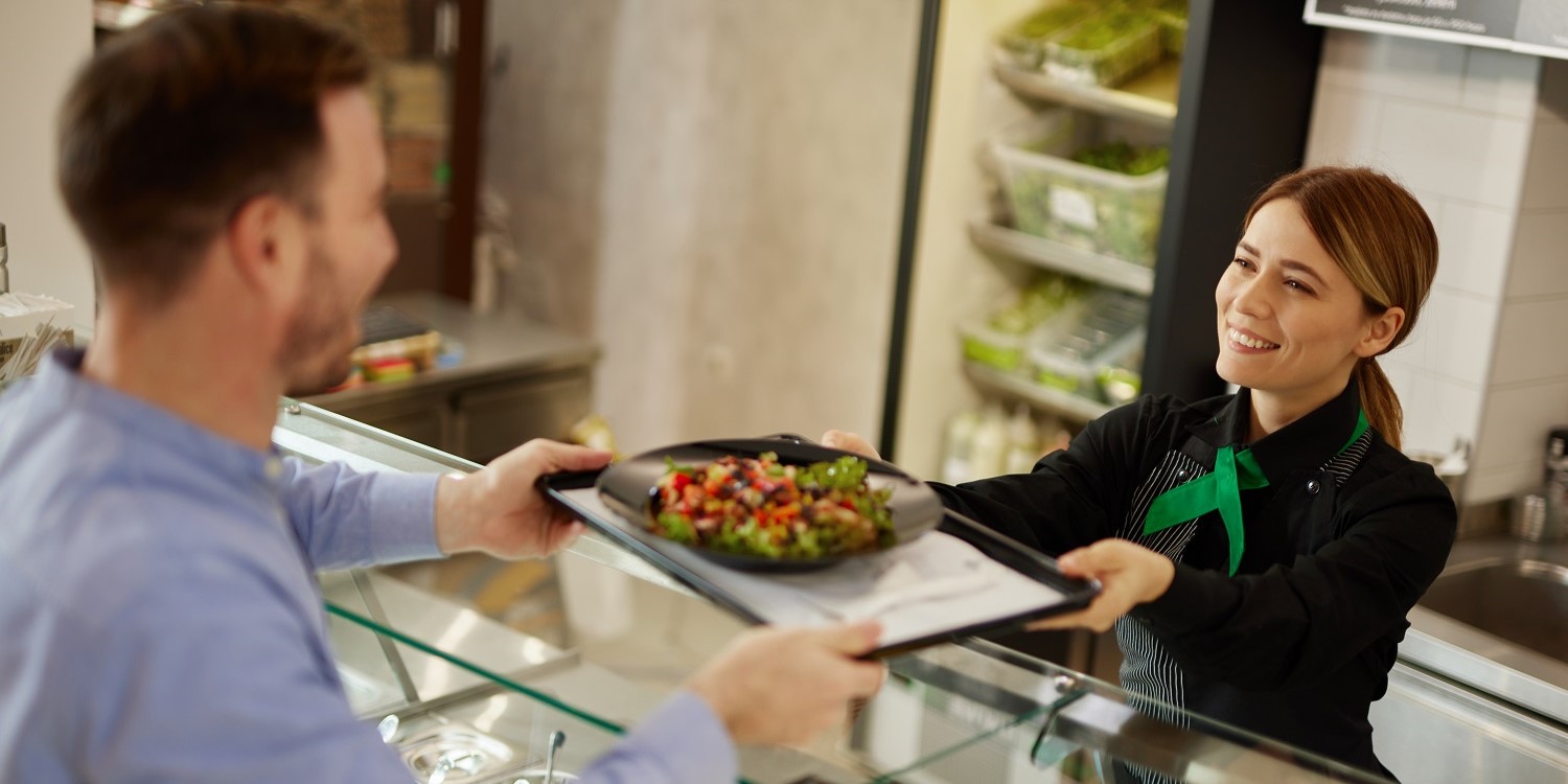 Eine Mitarbeiterin in der Kantine reicht einem Mann ein Tablett mit einem Gemüsegericht über die Theke. . Artikel "Alternativen zu Fleisch in Kantine und Co." öffnen.