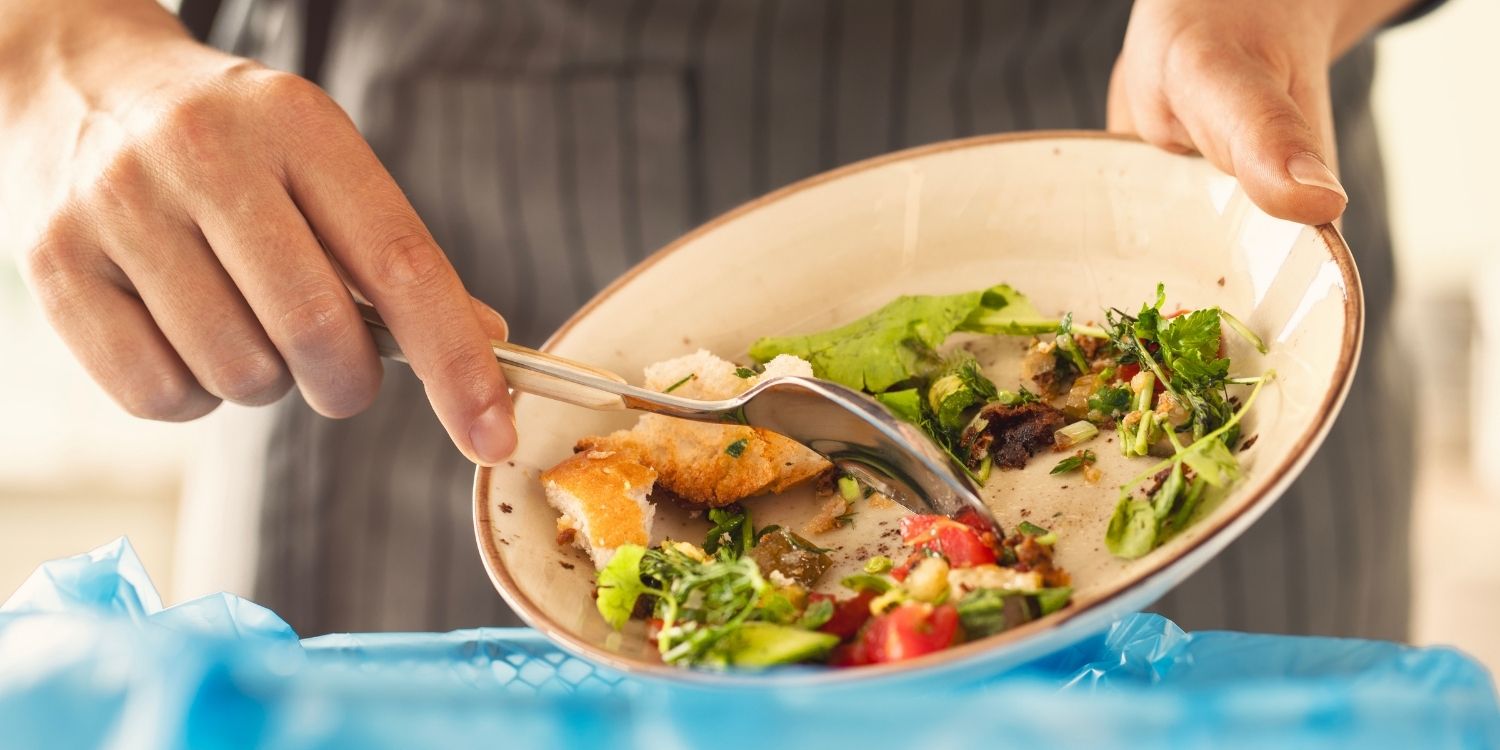 Eine Frau mit Schürze leert Essensreste von einem Teller in einen Mülleimer. . Artikel "Wählbare Portionen in der Gastronomie – leider kein Standard" öffnen.